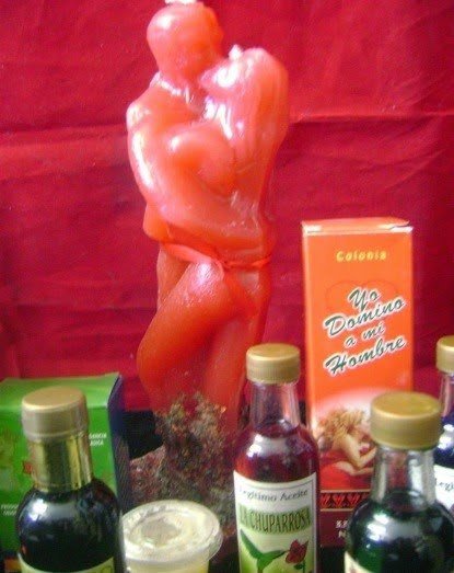 Vela roja con forma de pareja abrazada, atada con cinta, junto a frascos de aceites y colonia