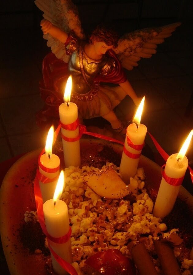 Figura del arcángel Miguel con velas blancas atadas con cinta roja alrededor de un cuenco con ofrendas