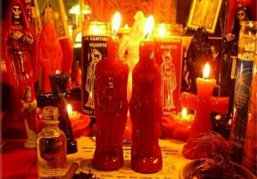 Dos velas rojas con figura humana encendidas en un altar con velas de la Santa Muerte, una cruz roja y frascos