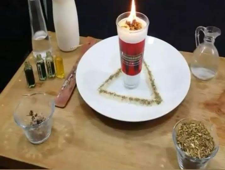 Vela roja y blanca encendida sobre un plato con un triángulo de hierbas, rodeada de frascos de aceites y hierbas