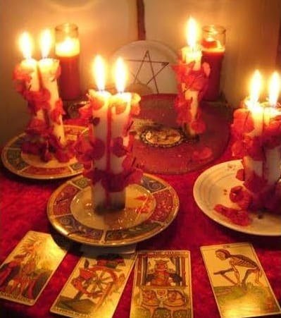 Velas blancas en pareja cubiertas de pétalos rojos, un pentáculo y cartas de tarot sobre un mantel rojo